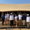 A group of smiling international travelers and local hospitality staff standing together in front of a luxury tented camp in the Serengeti.