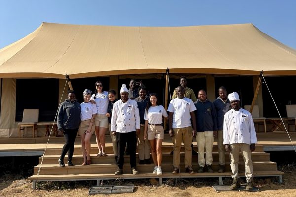 A group of smiling international travelers and local hospitality staff standing together in front of a luxury tented camp in the Serengeti.