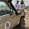 Two travelers wearing branded Gagago t-shirts sitting in an open-top safari jeep parked on the Serengeti plains.