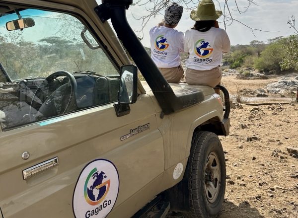 Two travelers wearing branded Gagago t-shirts sitting in an open-top safari jeep parked on the Serengeti plains.